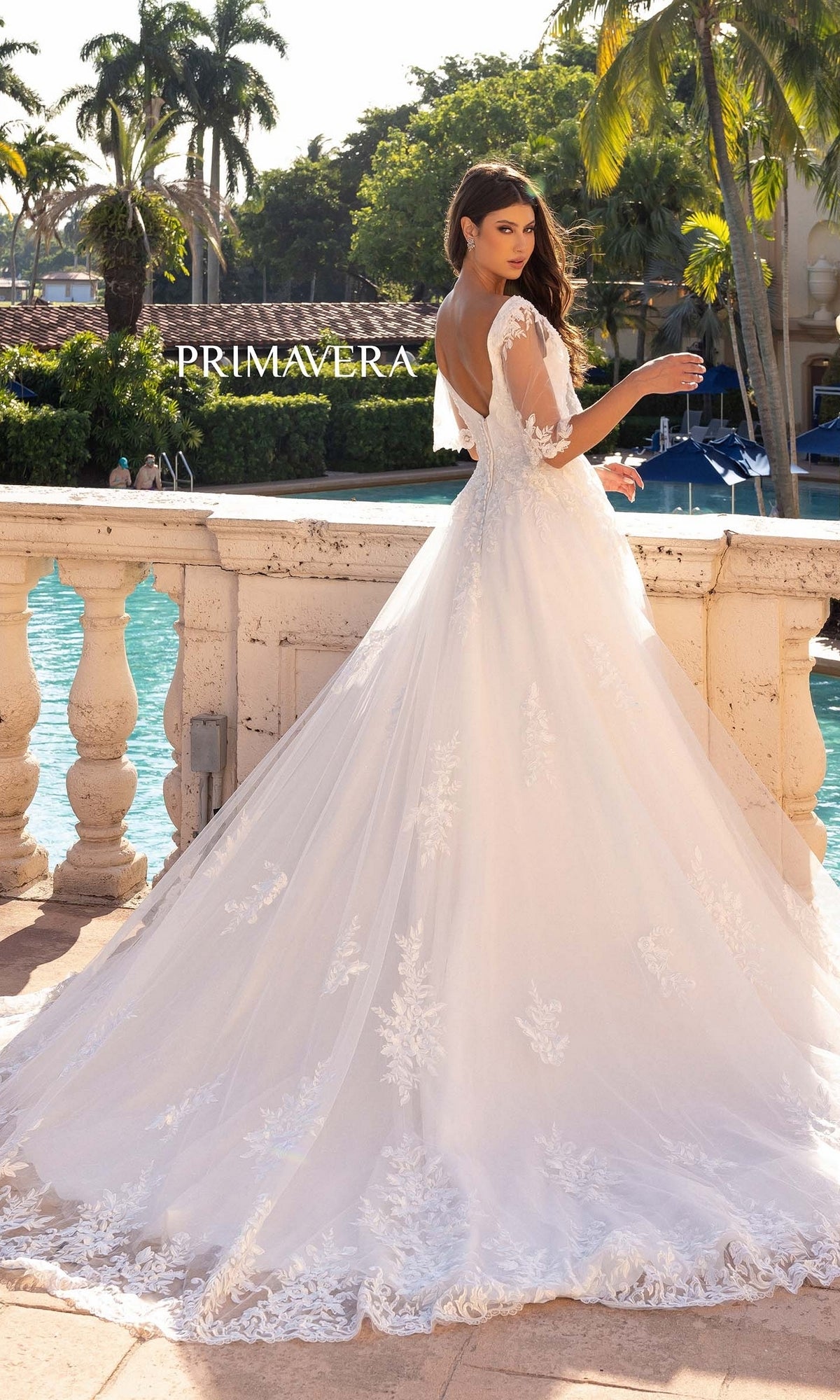 A woman in the Long Wedding Dress 11134 by Primavera—an ivory A-line gown with lace details and cape short sleeves—stands by a stone railing, overlooking a pool with palm trees and a